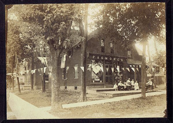 Store with name Pasmore over window and family sitting outside. There are pennants and flags decorating the trees around the building.