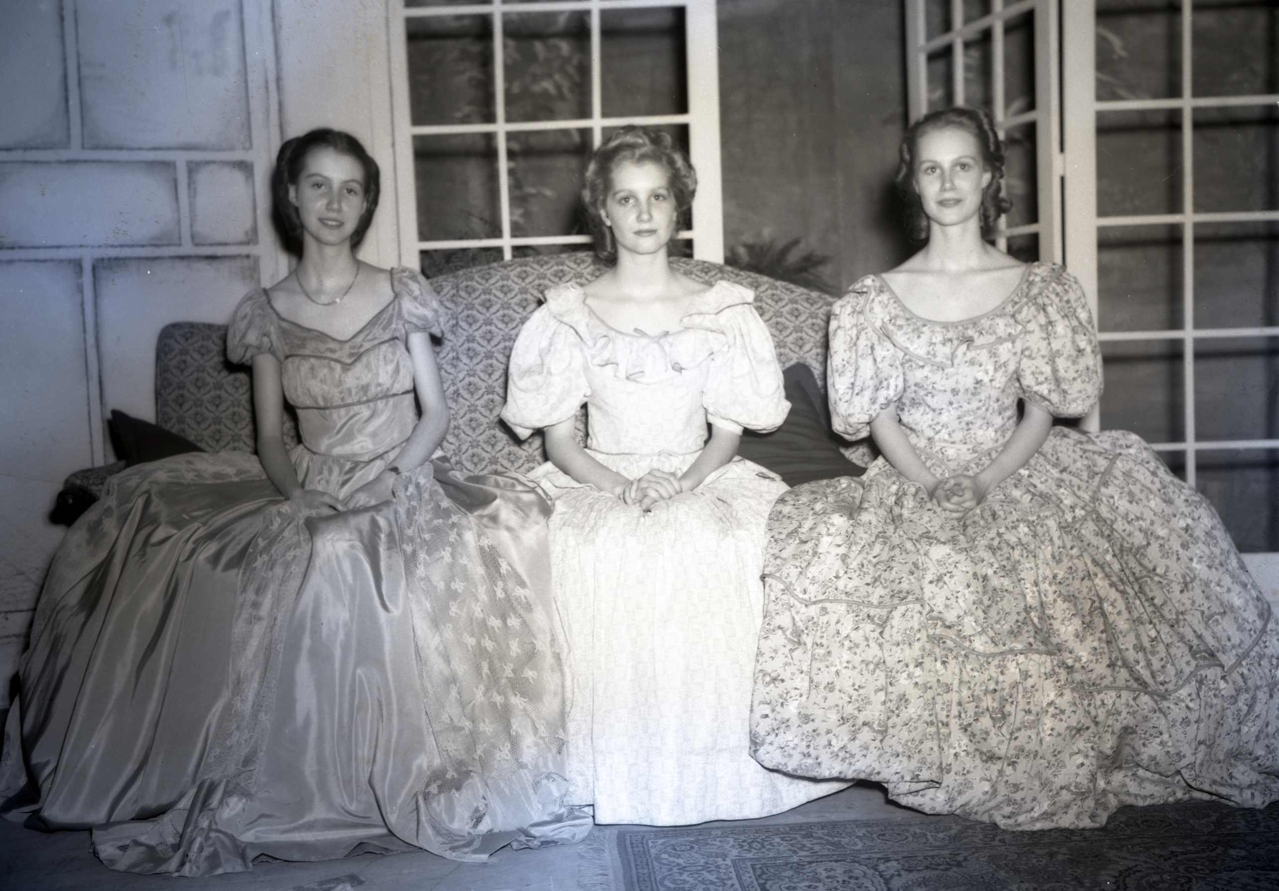 Three young women sitting on a chair wearing long dresses.