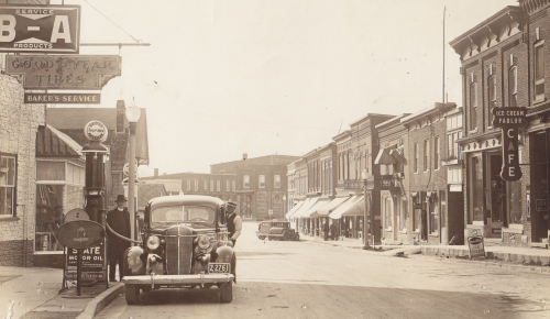 Man filling a 1920s car with gas on the street of a town.