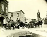 Firemen outside fire hall with motorized and horse-drawn vehicles.